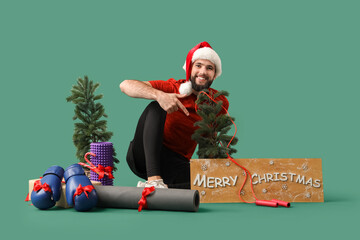 Young happy man in Santa hat with sport equipment, trees and text MERRY CHRISTMAS sitting on green background