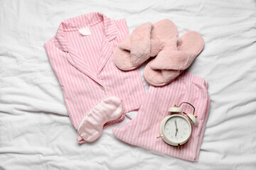 Stylish pajamas with slippers, sleep mask and alarm clock on white bed sheet