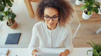 A woman with curly hair and glasses is sitting at desk, using tablet in bright, modern workspace filled with plants. She appears focused and engaged in her task