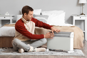 Fototapeta premium Young man warming hands near electric convector heater in bedroom