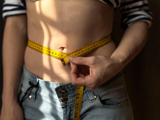 Close up of faceless woman in jeans measuring her waist with yellow tape measure. Weight loss, fitness and body measurement concept for health and wellness. Lifestyle photo.
