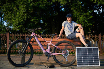 Man cyclist using solar panel for charging electric mountain bike outdoors. Guy in sunglasses sits...