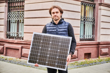 Man holding solar panel, stands in front of old building. Guy in a black hoodie and navy blue...