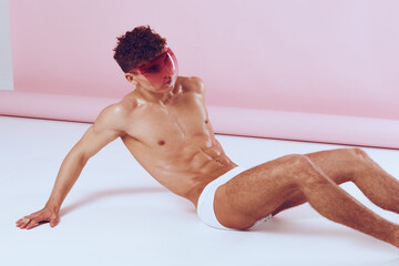 Stylish male model posing confidently in minimalistic pink studio, wearing bright sunglasses and white underwear. Fashion and self-expression concept.