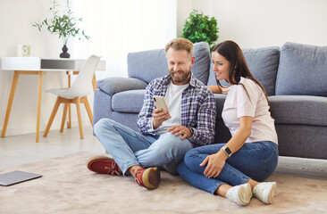 Portrait of happy smiling couple sitting on sofa in living room at home and using smartphone...
