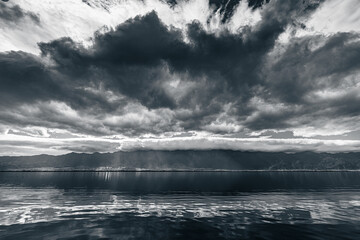 Fototapeta premium Dramatic monochrome landscape of Erhai Lake in Dali, Yunnan, China. Heavy clouds contrast with the serene water, reflecting the mountains and sky. Sunbeams pierce the clouds, creating a breathtaking