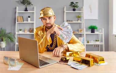 Portrait of a serious rich man in a gold suit holds cash money while working on a laptop. Wealth, prosperity, and financial success, highlighting his dedication and serious approach to his work.