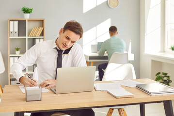 Obraz premium Man businessman working in an office with a laptop, engaged in a phone call with a client. Professional communication and multitasking, businessman and client or customer conversation.