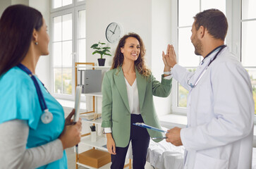 Obraz premium Friendly doctor with nurse rejoicing at success in treatment giving high five to smiling woman patient standing in clinic after medical examination. Health care and medicine concept.