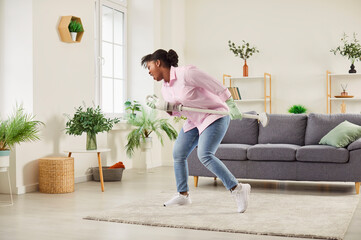 Obraz premium Happy excited funny middle aged african american woman singing at vacuum cleaner hoover cordless tube as microphone, cleaning floor carpet at home, enjoying household activities, vacuuming, having fun