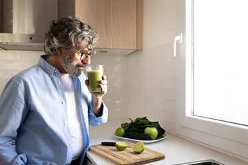Mature man drinking healthy vegetable green smoothie for breakfast standing in kitchen. Copy space.