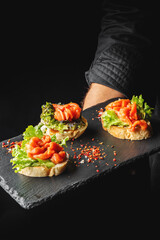 A close-up of an elegant platter featuring three gourmet canapés topped with fresh salmon, lettuce, and red pepper flakes, served on a black slate board. Ideal for food and culinary themes.