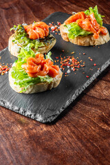 Three delicious open-faced sandwiches topped with fresh salmon and crisp lettuce, served on a black slate board. A sprinkle of red chili flakes adds a touch of spice to this appealing appetizer.