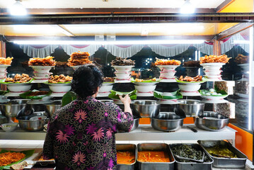 A man serving customers, Indonesian food, Padang cuisine buffet: Fish in coconut milk, fried fish, grilled fish, beef, and other dishes are displayed in a glass display case.