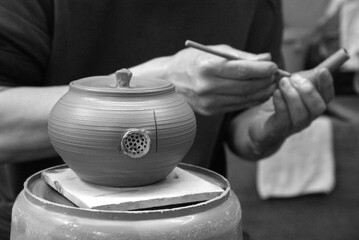 A potter adds fine details to a clay teapot "kyusu", crafting a strainer by hand. This careful work highlights Japan’s mastery of functional yet elegant pottery techniques. Black and white image.