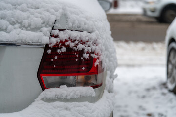 Close-up rear view or elegant red tail light of modern white coloured car covered with fresh white snow outdoors on winter city street. Soft focus. Copy space. Automobile details theme.