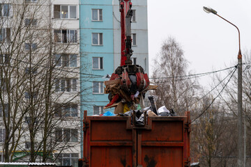 Fototapeta premium Side view of orange machine crane loader manipulator with crab bucket holding bunch of garbage over garbage truck in urban residential district in a cloudy autumn day. Soft focus. Industrial machines.