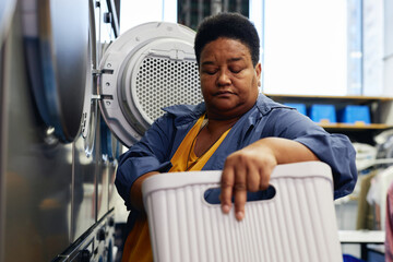 Medium portrait shot of senior Black woman loading clothes from basket into washing machine organizing daily laundry at self service laundromat