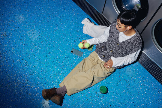 Top view of young man listening to music in headphones while passing time in self service laundry sitting on floor and playing with tennis balls in hand, camera flash, copy space