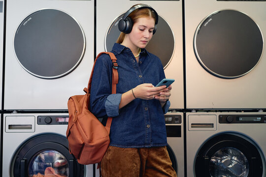 Medium full shot of young woman in casual outfit enjoying music in headphones text messaging on phone, while waiting in coin laundromat with washing machines tumbling in background