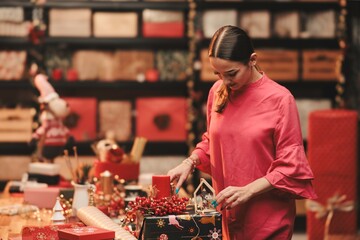 A woman in a vibrant pink blouse focuses intently on crafting festive decorations in a warm and inviting holiday workshop, surrounded by cheerful decor and handmade treasures