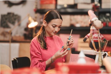 Inside a warmly decorated space, a woman enjoys the festive spirit while meticulously painting Christmas ornaments. Surrounded by colorful supplies, she immerses herself in creative bliss