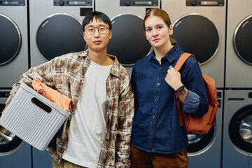 Portrait shot of young woman and Asian man with basket of clothes posing against washing machines...