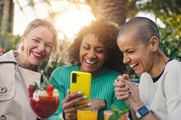 Fototapeta premium Three cheerful multi-ethnic female friends enjoying drinks at a bar, having fun watching something on a mobile phone app