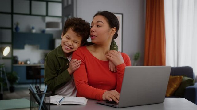 Busy concentrated African American business woman mother businesswoman freelancer female girl using laptop working computer online at home loving child boy kid son coming hugging mom cuddling family