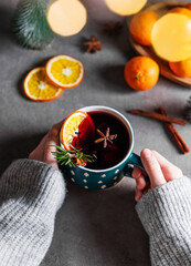 Hands in a sweater holding a cup of mulled wine with orange, cinnamon and rosemary on a dark background