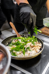 A chef in a black glove adds fresh herbs to a skillet with cooked fish, enhancing the dish's flavor. The vibrant ingredients showcase a focus on culinary art in a professional kitchen setting.