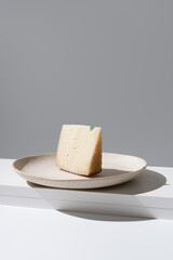Trozo de queso de oveja curado. Cuña de queso manchego en un plato beige sobre mesa blanca y fondo gris	