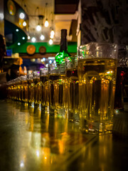 Golden Glow of Celebration. A row of golden-hued glasses aligned on a table in a warmly lit bar, capturing the lively ambiance with blurred lights and shadows in the background.