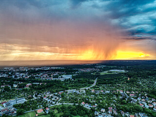 Sunset Rainstorm Over Polish City. A dramatic aerial view of a sunset rainstorm over a Polish city, with vivid orange and yellow skies contrasting against dark clouds and urban greenery.