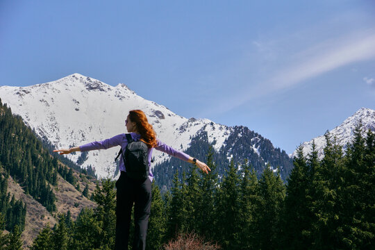 A young girl stands with her arms outstretched against the background of snowy mountain peaks in a mountain valley on a sunny day - Powered by Adobe