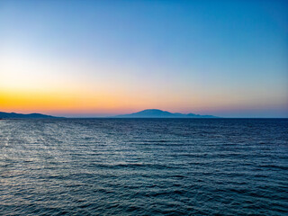 Distant Mountain Sunset. A calm ocean stretches to the horizon as the sunset casts warm hues over distant mountains, blending seamlessly into the tranquil sky.