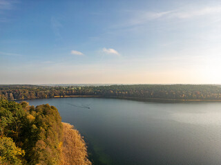 Obraz premium Peaceful Lake with Autumn Forest Horizon. A serene aerial view of a calm lake surrounded by dense autumn forests, with golden sunlight illuminating the tranquil landscape under a clear blue sky.