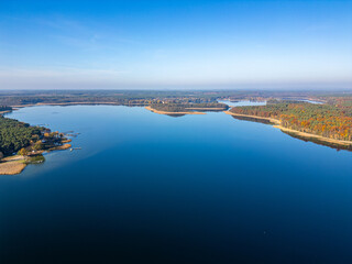 A sweeping aerial view of a vast lake surrounded by lush forests with docks and tranquil waters, framed by a clear blue sky.