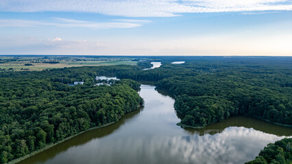 Fototapeta premium Serene Forested Lake Raduń in Wałcz, Poland. Aerial view of Lake Raduń winding through dense forests, with calm reflective waters and expansive rural fields stretching toward the horizon in Wałcz