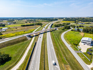 Highway Overpass with Rural Backdrop. Aerial view of a highway intersected by an overpass, surrounded by green fields, rural buildings, and vibrant yellow patches under a clear blue sky.