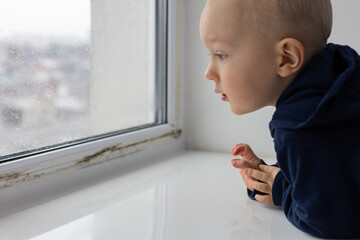 Little child looking through the window with water drops condensate and black spots of mouldy...