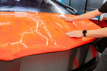 Person is smoothing out an orange vinyl wrap on car hood in workshop, ensuring bubble free finish for protect body automotive