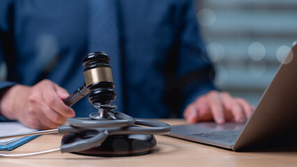 A man is sitting at a desk with a gavel and a laptop. He is typing on the laptop while holding the gavel. The scene suggests that the man is a lawyer or a judge who is working on a case