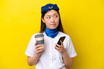 Airplane Chinese woman stewardess isolated on yellow background holding coffee to take away and a mobile while thinking something