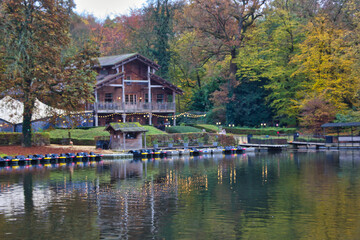 Fototapeta premium Beautiful autumn landscape in the park: chalet on the shore of the lake, with tiny boats anchored in front of it.