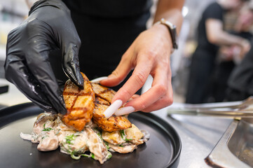 A chef's hand, wearing a black glove, carefully arranges grilled chicken pieces on a plate with creamy mushroom sauce. The kitchen setting is busy and vibrant, showcasing culinary artistry.