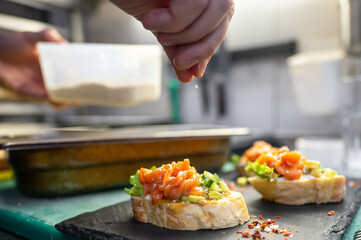 A chef sprinkles seasoning over gourmet bruschetta topped with fresh salmon and vegetables, showcasing culinary preparation in a modern kitchen. Close-up view highlights the art of food presentation.