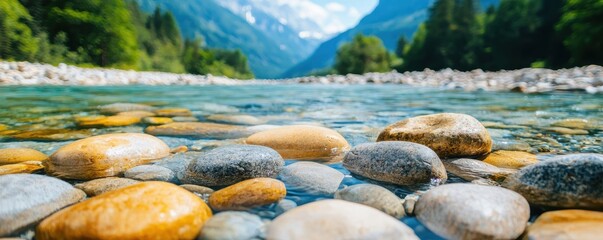 Fototapeta premium A serene river landscape featuring smooth stones, clear water, and lush greenery, framed by majestic mountains under a bright blue sky.