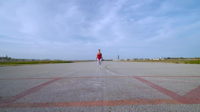 Una bailarina de danza cl&aacute;sica llega desde el infinito por una pista de aterrizaje de un aeropuerto y comienza a bailar un d&iacute;a soleado con cielo azul y nubes.