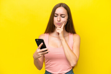 Young caucasian woman isolated on yellow background using mobile phone and thinking